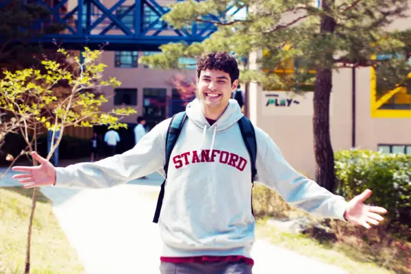 Noah Centineo as Peter Kavinsky, standing outside, wearing a Stanford jumper, with his arms outstretched and smiling