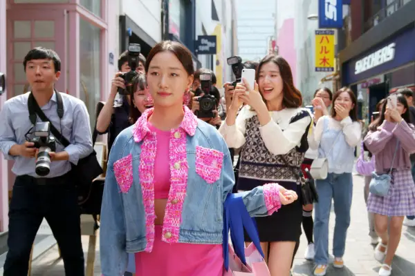 Yuri dressed in pink and denim walking down the street with a crowd following her and taking photos of her