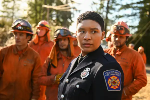 Jules Latimer as Eve Edwards in Fire Country season 2 standing in her black uniform while standing in front of a group of people wearing orange fire safety uniforms standing in the middle of a forest.