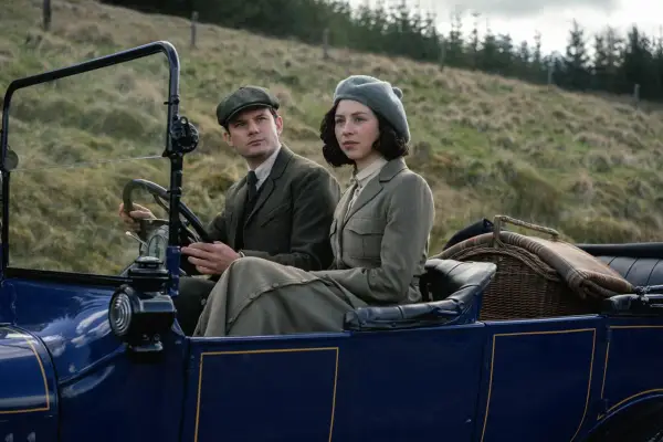 Jeremy Irvine as Henry Beauchamp and Hermione Corfield as Julia Moriston in Outlander: Blood of My Blood sitting in an open top car and looking nervous while driving in the countryside.
