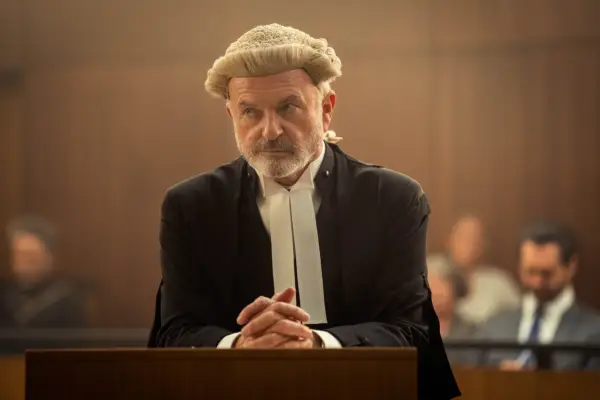 Sam Neill as Colby SC resting his hands against a wooden stand, wearing a barrister gown and wig in The Twelve.
