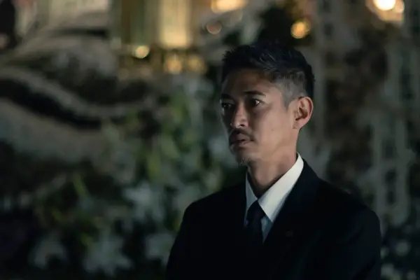 Hayama in an outdoor nighttime setting, with greenery behind him, wearing a white shirt and tie with a black coat