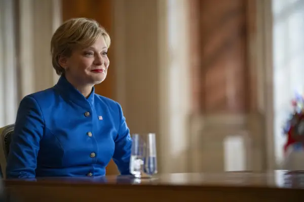Martha Plimpton as Senator Judith Holt in The Regime, wearing a bright blue jacket and smiling while sat down