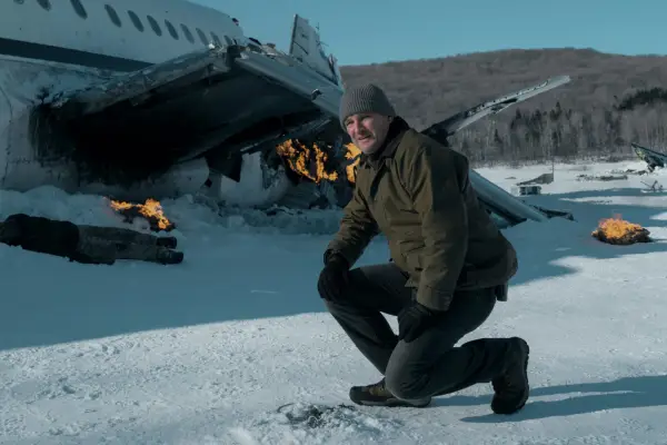 Jason Clarke stars in The Last Frontier; his character is kneeling down in a snowy wilderness area, with a crashed plane in the background behind him