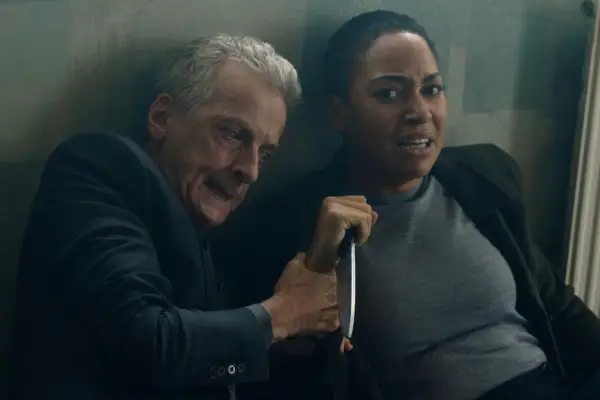 Cush Jumbo and Peter Capaldi sat down next to each other with looks of anguish. Jumbo is holding a knife and looks scared while Capaldi his holding her hand