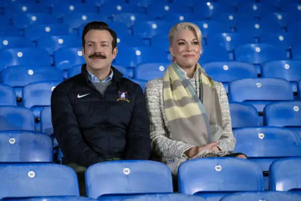 Jason Sudeikis and Hannah Waddingham in Ted Lasso. They are sat in a football stadium and are smiling