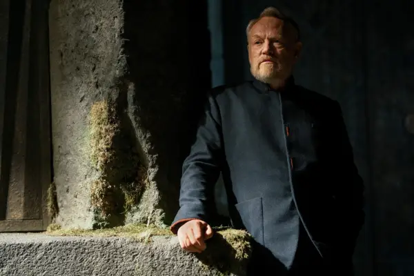 Jared Harris stars in Foundation season 3. His character is leaning against a stone pillar in a dark, shadowy environment