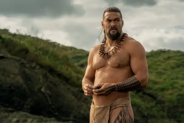 Jason Momoa stars in Chief of War; he is seen here shirtless against a cloudy backdrop with large green hills, looking stern and deep in thought
