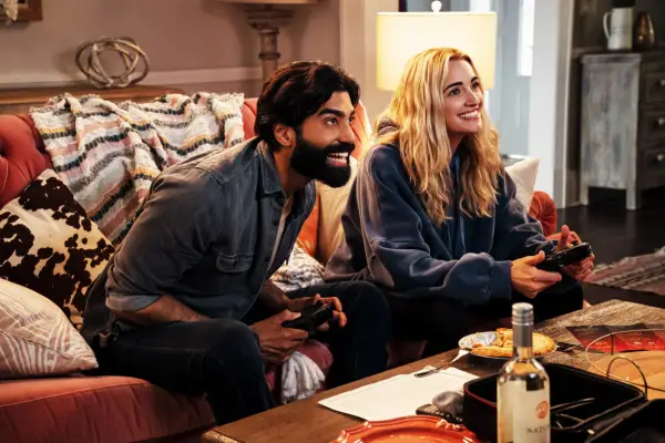 Raymond Ablack as Joe and Brianne Howey as Georgia, sat next to one another on a sofa playing a video game