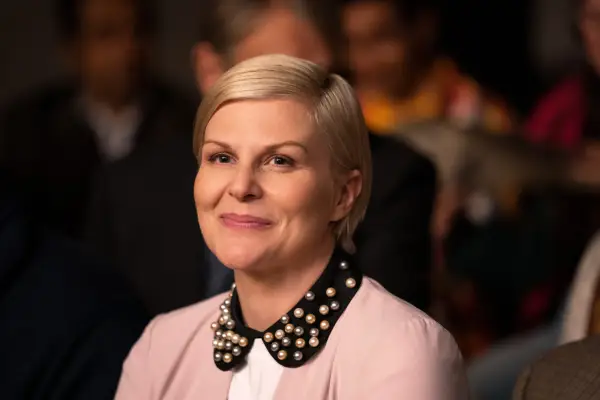 Jennifer Robertson as Ellen, smiling, sat in an audience, wearing a pink top with a black collar