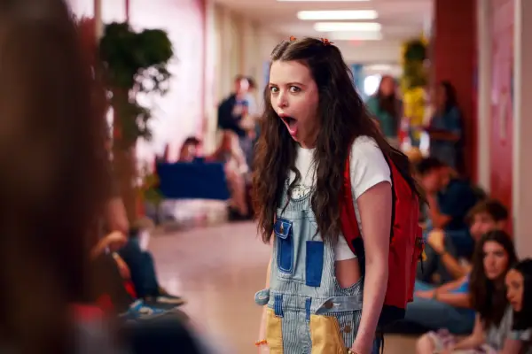 Sara Waisglass as Maxine, standing in the school corridor, wearing dungarees, wide wide open in shock