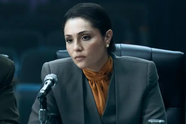 June dressed in a grey suit, sat at a desk in a court room, with a microphone in front of her