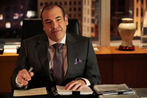Rick Hoffman as Louis Litt in Suits sat at a desk with a pen in his hand