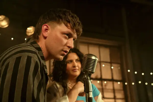 Tom Francis as Clayton, Natasha Behnam as Dominique in You season 5 standing on stage and speaking into a microphone, looking into the crowd of people.