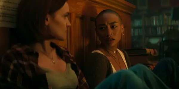 Tati Gabrielle as Marienne Bellamy sits on the right of Madeline Brewer as Bronte in a dimly lit room in You season 5.