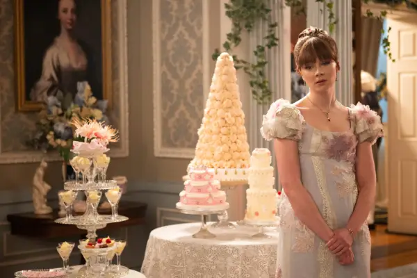 Claudia Jessie as Eloise Bridgerton in Bridgerton standing in front of a white wedding cake and decorations, looking sad as she wears a ballgown.
