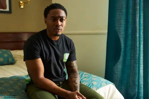 Ben sits on a hotel room bed, with a troubled expression on his face