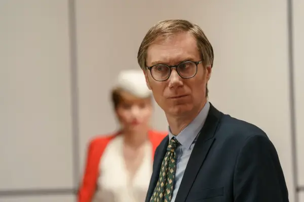 Greg (Stephen Merchant) looks worriedly at someone in a courtroom, with Lady Gabby (Eleanor Tomlinson) standing out of focus behind him