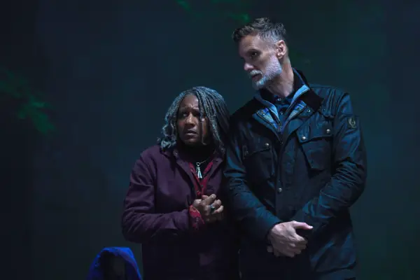 Myrna stands close to John, looking concerned on a dark night in the forest