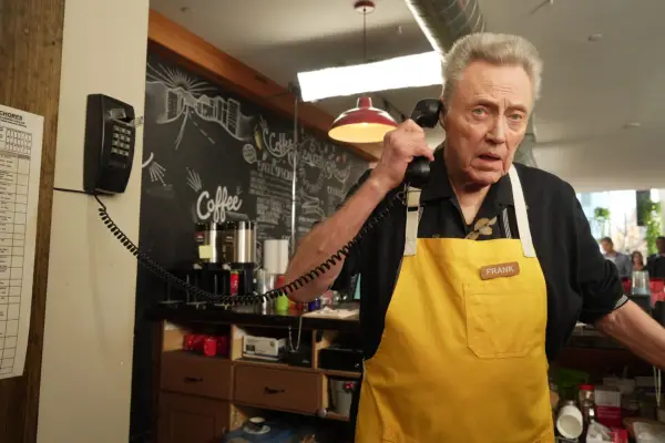 Frank (Christopher Walken) wearing a yellow apron while working in a cafe, holding a wired landline phone to his ear