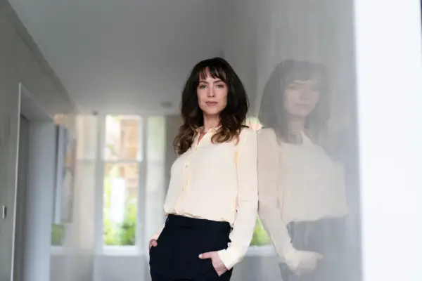 Rhona standing in a white hallway, leaning against the wall