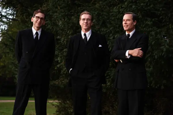 Nicholas, Kim and Angleton stood in a garden in a line, all wearing black suits