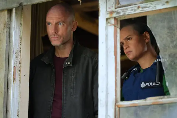 Richard Flood as Theo Richter and Acushla-Tara Kupe as Diana Huia in The Gone season 2 both standing inside a house and staring out of the open window with serious expressions.