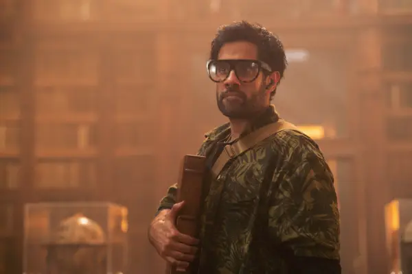 Sacha Dhawan in The Iris Affair, holding a book and wearing glasses.