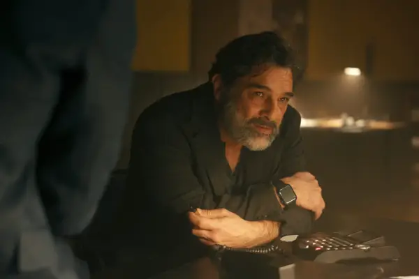 Marco Leonardi as Bruni in The Iris Affair, wearing a black shirt and sat at a desk with his arms crossed.