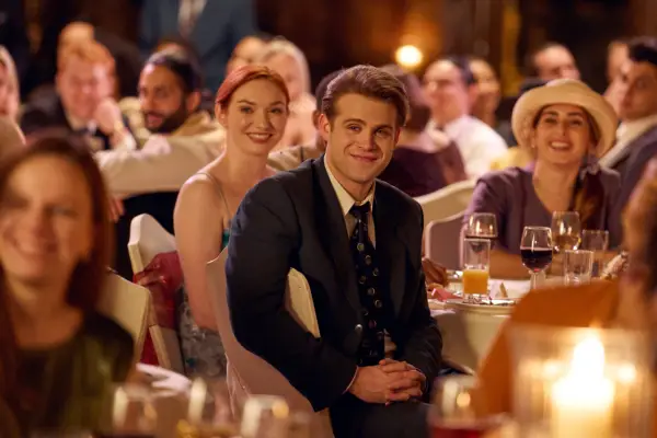 Leo Woodall as Dexter Mayhew in One Day sitting in a hall at a table with others but turned around and staring at the stage.