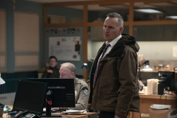 Christopher Eccleston wears a suit and coat in a police station in a scene from True Detective: Night Country