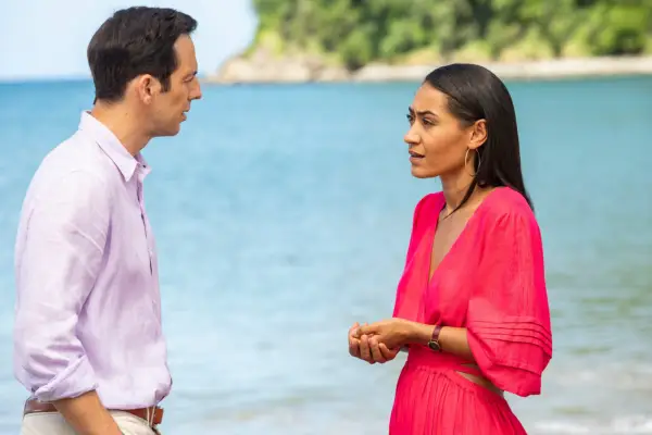 Ralf Little as Neville Parker and Joséphine Jobert as Florence in Death in Paradise. They are talking on the beach with the sea in the background.