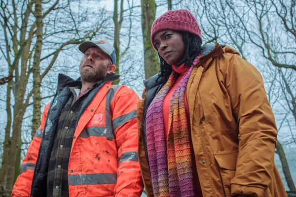 Wunmi Mosaku as DC Riya Ajunwa and Hubert Hanowicz as Jakob Makowski in Passenger standing in the middle of a snowy forest and looking down at something.
