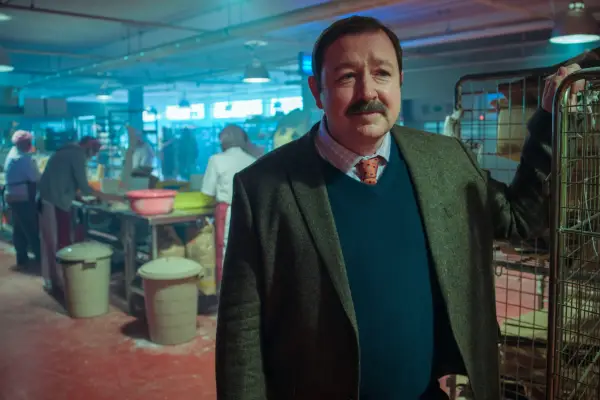 Daniel Ryan as Derek Jackson standing in the middle of a factory wearing a suit and smiling in Passenger.