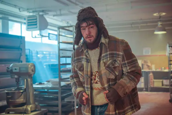 Nico Mirallegro as Kane Jackson looking scared standing in the middle of a factory wearing a coat and hat in Passenger.