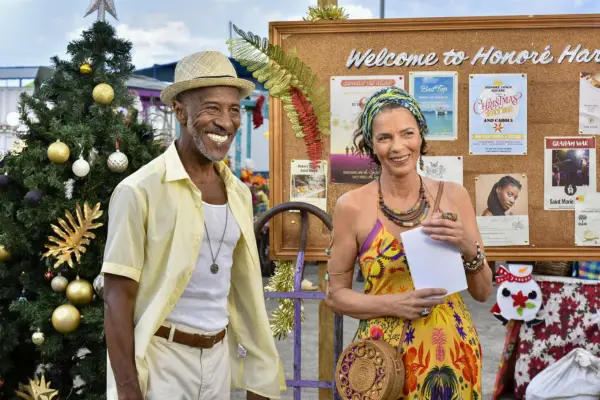 Danny John-Jules as Dwayne Myers and Elizabeth Bourgine as Catherine Bordey, standing next to one another smiling with the welcome to Saint Marie board and a Christmas tree behind them