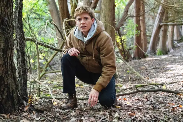 Mae Martin as Alex Dempsey, crouching down in a forest, wearing a warm jacket and dark trousers.