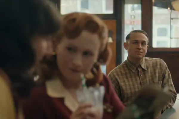 Charlie Hunnam as Ed Gein in Monster: The Ed Gein Story sitting on a stool at a counter, watching two women have a conversation.