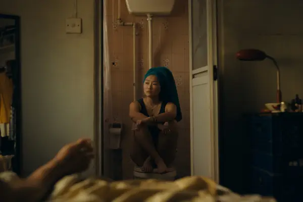 Ji-young Yoo as Mercy sitting cross legged on a toilet seat with a towel wrapped around her head, facing a man lying on her bed.