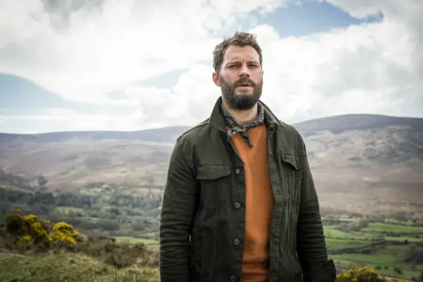 Jamie Dornan in The Tourist wearing an orange jumper and green jacket, with fields in the background