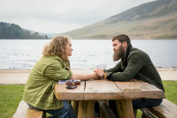 Helen (Danielle Macdonald) and Elliot (Jamie Dornan) in The Tourist holding hands, sitting on a wooden bench by a lake