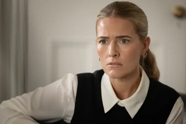 Amy De Bhrún in Borderline sitting on a sofa, wearing a white shirt and black jumper, looking worried.
