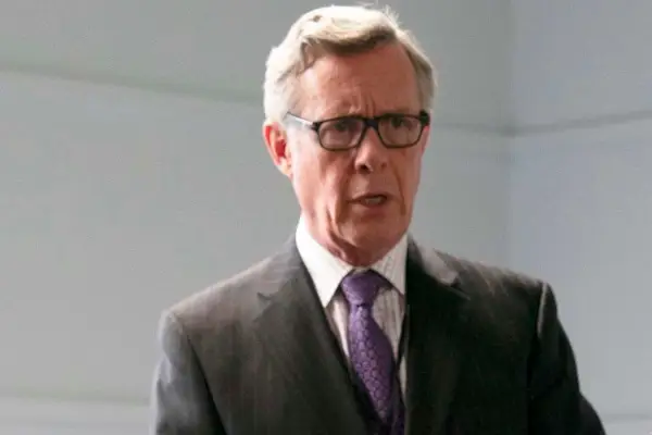 Alex Jennings as James Arbuthnot, wearing a suit, stood up talking to people