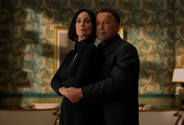 Carrie-Anne Moss and Arnold Schwarzenegger standing in an ornate room and embracing, as if in the middle of a dance routine, with both of them wearing black.