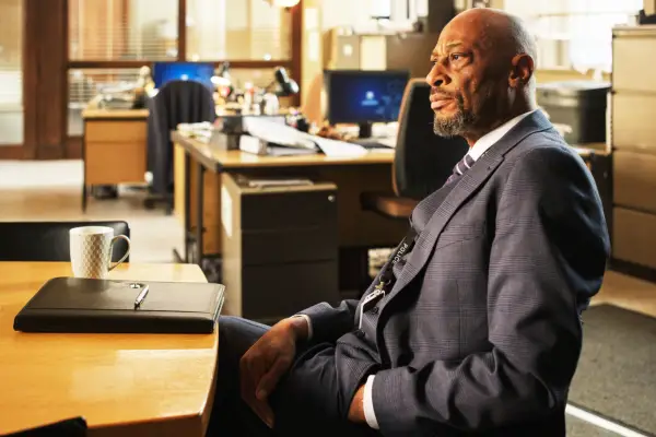 Terence Maynard plays DCI Paul Goodwin, wearing a suit, sat behind a desk in an office