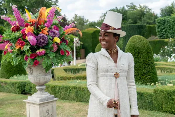 Adjoa Andoh as Lady Agatha Danbury in Bridgerton wearing an all white suit and hat, smiling while holding a walking stick and in the garden.