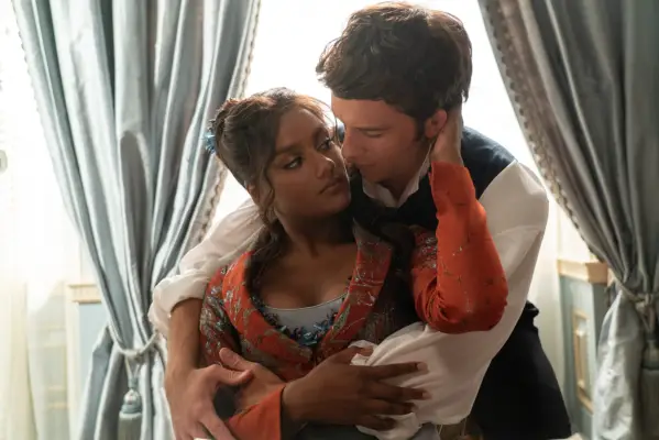 Simone Ashley and Jonathan Bailey holding each other in an embrace in front of a window.