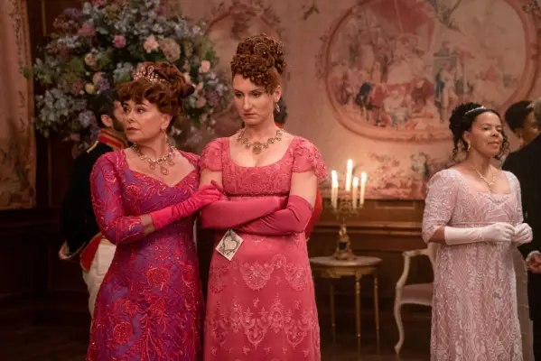 Polly Walker as Lady Portia Featherington, Bessie Carter as Prudence Featherington standing to the side of a ball both wearing pink and looking stressed in Bridgerton.