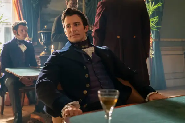 Luke Thompson as Benedict Bridgerton leaning back in his chair in the middle of a bar and looking intrigued by something in Bridgerton.