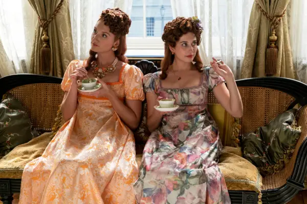 Bessie Carter as Prudence Featherington, Harriet Cains as Philipa Featherington sitting on a sofa and drinking tea from a cup and saucer in Bridgerton.